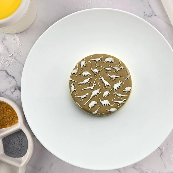 Decorative cookie with dinosaur pattern on a white plate surrounded by baking tools on a marble surface