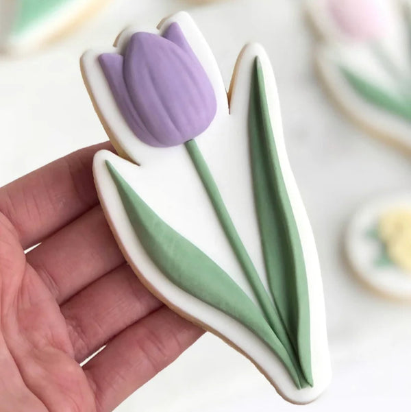 Decorative cookie shaped like a tulip with purple flower and green leaves held in a hand.